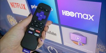 Roku remote in hand in front of a Roku home screen.