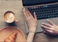 Hands on a MacBook keyboard alongside a coffee and croissant.