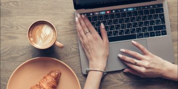 Hands on a MacBook keyboard alongside a coffee and croissant.