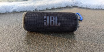 A JBL Flip 6 Bluetooth speaker sitting on wet sand by the ocean.