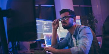Profile photo of a developer / programmer reviewing code on monitors in his workspace.