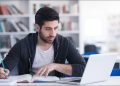 Young man studying in a library and using a laptop.