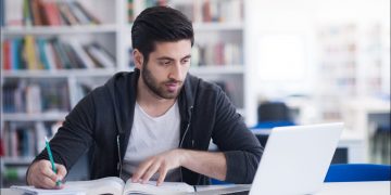 Young man studying in a library and using a laptop.