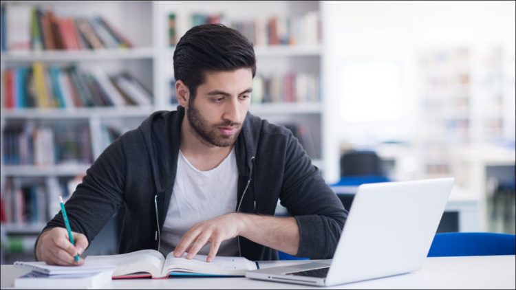 Young man studying in a library and using a laptop.