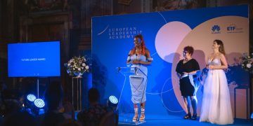 A young woman stands on stage holding a glass award while speaking.