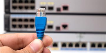 A person's hand holding an Ethernet cable connector in front of internet switches.