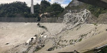 A site of debris with trees and a cloudy sky in the background. It is the Arecibo Observatory after it collapsed in 2020.