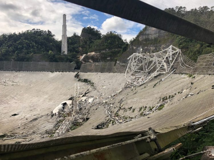 A site of debris with trees and a cloudy sky in the background. It is the Arecibo Observatory after it collapsed in 2020.